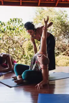 A yoga teacher assists a participant in a seated spinal stretch on a yoga mat, while other participants practice nearby on a wooden floor surrounded by tropical greenery.