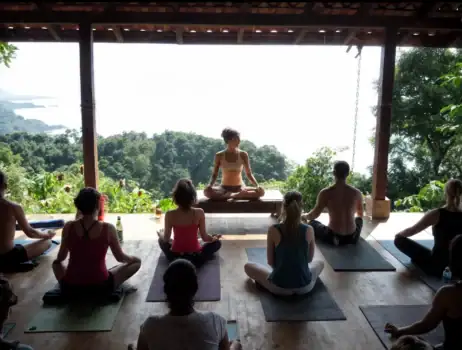 The image depicts a yoga class taking place in an outdoor pavilion with a scenic view of lush greenery and trees. The participants are sitting on yoga mats, and the instructor is l